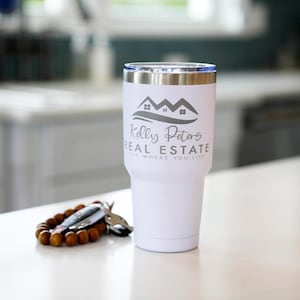 May include: A white insulated tumbler with a silver rim and a blue lid. The tumbler features the text "Kelly Peters Real Estate" and a house logo. A wooden beaded bracelet and car keys are next to the tumbler.