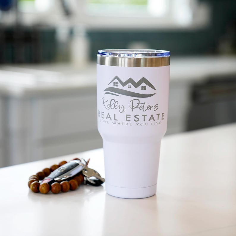 May include: A white insulated tumbler with a silver rim and a blue lid. The tumbler features the text "Kelly Peters Real Estate" and a house logo. A wooden beaded bracelet and car keys are next to the tumbler.