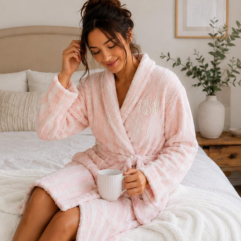 May include: A light pink and white striped plush robe with a shawl collar and a matching belt. The robe has embroidered initials on the chest. A white mug is held in the lap. The robe is on a bed with white bedding.