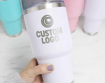 May include: A white stainless steel tumbler with a metallic rim and a clear lid, featuring a circular graphic and the text CUSTOM LOGO printed in gray. The tumbler is held in the foreground with other colorful tumblers visible in the background.
