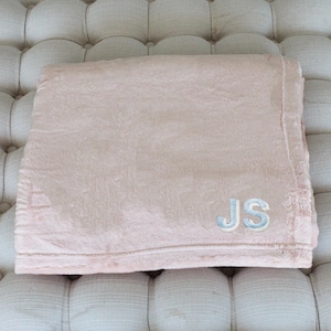 May include: A folded, blush-colored blanket with a soft, plush texture. The blanket features the embroidered initials "JS" in light blue thread, adding a personalized touch. The blanket is a cozy home textile.