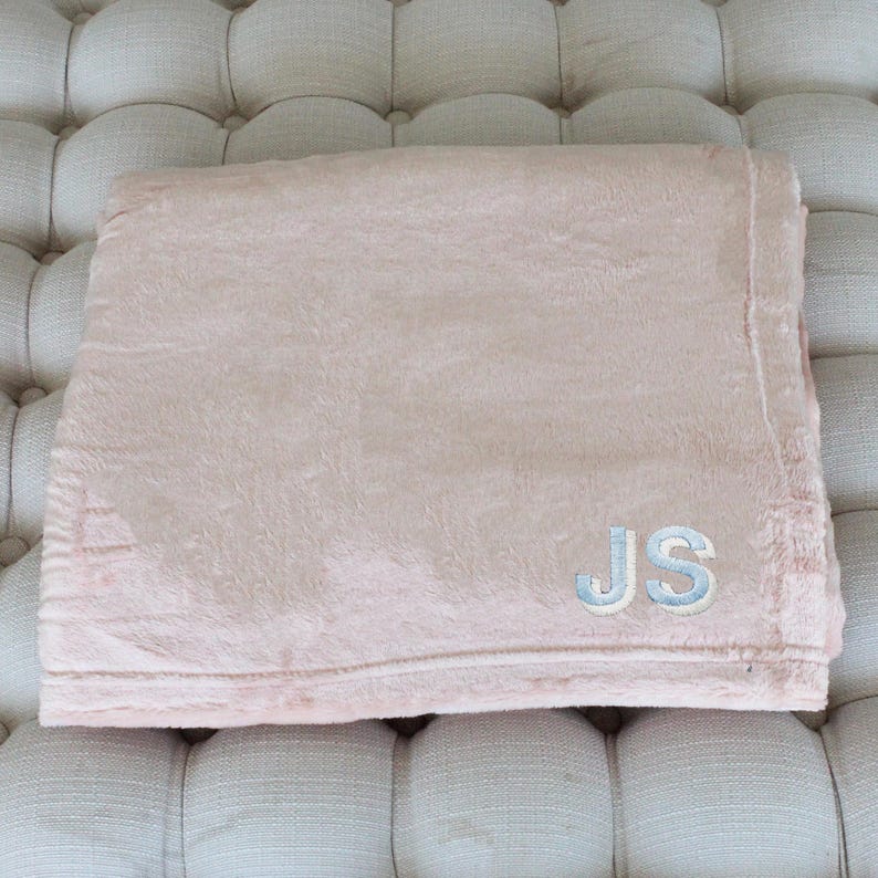 May include: A folded, blush-colored blanket with a soft, plush texture. The blanket features the embroidered initials "JS" in light blue thread, adding a personalized touch. The blanket is a cozy home textile.