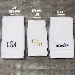 May include: Three white terry cloth towels with embroidery. The left towel features a charcoal serif monogram "GSM". The center towel displays a gold diagonal line monogram "C/M". The right towel shows a navy block font name "brooks".