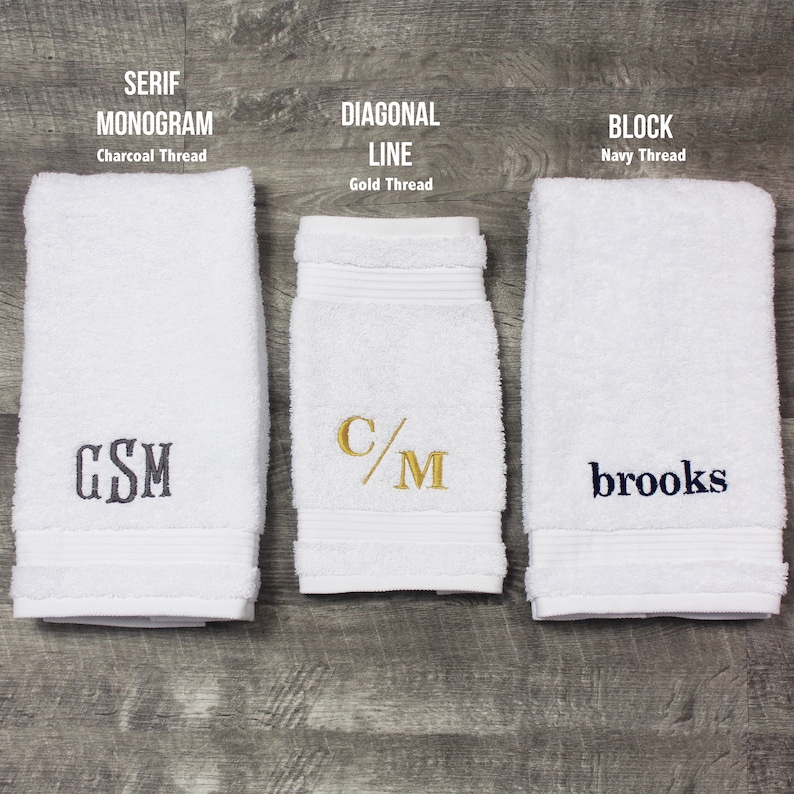 May include: Three white terry cloth towels with embroidery. The left towel features a charcoal serif monogram "GSM". The center towel displays a gold diagonal line monogram "C/M". The right towel shows a navy block font name "brooks".