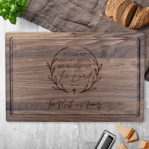 May include: A rectangular wooden cutting board with a dark brown finish. The board features engraved text within a decorative wreath: "AS FOR ME AND MY HOUSE WE WILL SERVE The Lord" and "The Matthews Family". A groove runs around the edge.