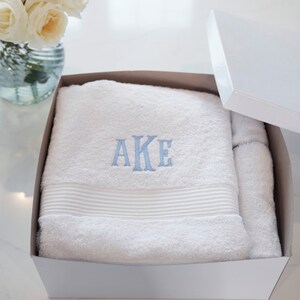 May include: A white cotton towel folded inside a white gift box. The towel features a textured ribbed border and the letters AKE embroidered in light blue thread in the center. The fabric has a soft, plush pile texture throughout the surface.