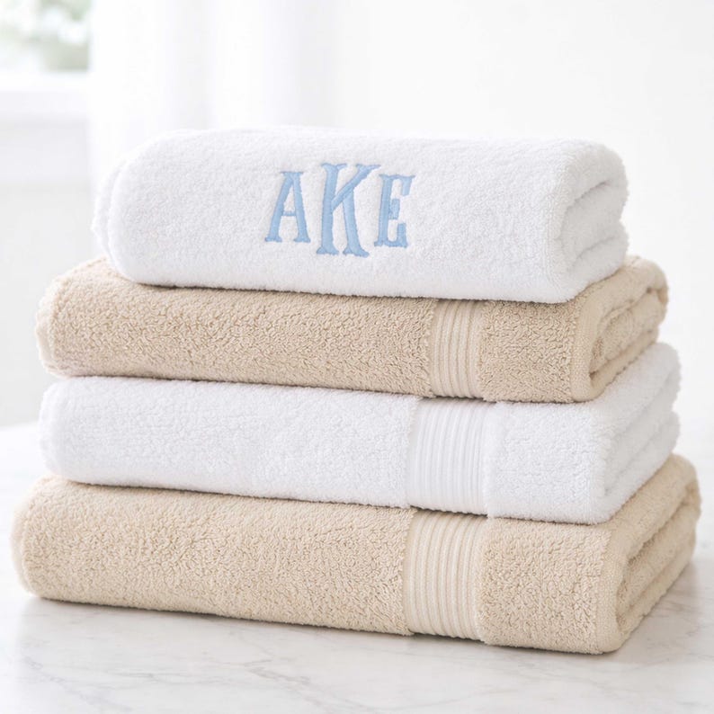 May include: A stack of four folded towels in alternating white and beige colors. The top white towel features the embroidered blue letters AKE in a serif font. Each towel has a textured terry cloth surface and a ribbed border detail.
