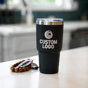 May include: A black insulated tumbler with a silver rim and a clear lid. The tumbler has the words "CUSTOM LOGO" printed in white. A wooden beaded bracelet and keys are next to the tumbler.