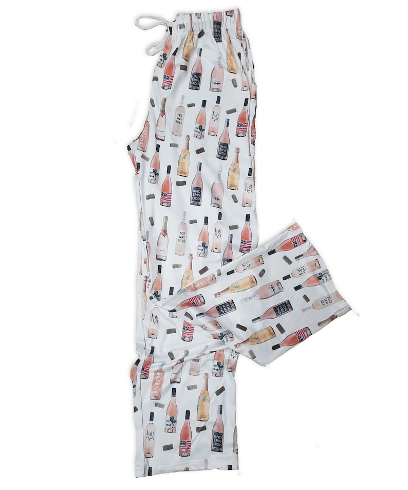 May include: White pajama pants with a pattern of pink wine bottles. The bottles have various labels with text such as "Yes Way Ros&eacute;" and "Summer Water".