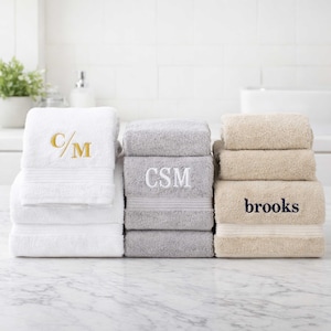 May include: A stack of plush cotton towels in white, gray, and beige. The white towel features gold embroidered initials C/M, the gray towel has white embroidered initials CSM, and the beige towel displays the name brooks in dark blue embroidery.