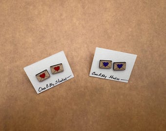 May include: Wooden envelope stud earrings with red and purple heart details. The earrings are displayed on white cards with the text "One fifty Studios" in black script. The background is a light brown surface.