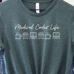 May include: A heather forest green t-shirt featuring the words "Medical Coder Life" in white script, along with coffee cup and laptop graphics. The shirt is displayed on a wooden hanger, with a denim jacket and a brown bag visible in the background.