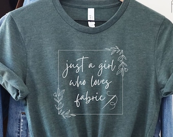 Op de afbeelding: Heather forest groen t-shirt met de tekst "just a girl who loves fabric" in een witte vierkant met bloemaccenten. Het shirt wordt getoond op een hanger met een spijkerjasje en een bruine leren tas op de achtergrond.