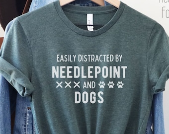 Op de afbeelding: Een groen T-shirt met de tekst "Easily Distracted by Needlepoint and Dogs" in wit. De tekst wordt omringd door drie witte X'en en drie witte poot afdrukken.
