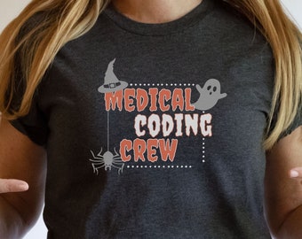 May include: Dark heather grey t-shirt with a Halloween-themed design. The design features a witch's hat, a spider, a ghost, and the text "Medical Coding Crew".
