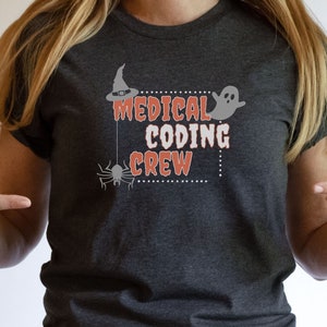 Puede incluir: Camiseta gris oscuro jaspeada con un diseño de temática de Halloween. El diseño presenta un sombrero de bruja, una araña, un fantasma y el texto "Medical Coding Crew".