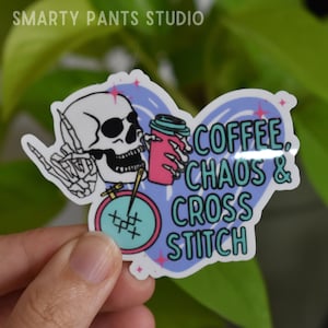May include: A sticker with a cartoon skull holding a cup of coffee and a needle and thread. The skull is wearing sunglasses and has a rock and roll hand gesture. The sticker says "Coffee, Chaos & Cross Stitch".