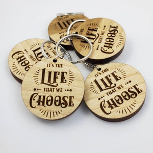 May include: Five wooden keychains with a laser-engraved design. Each keychain features the text "It's the life that we choose" with a stylized font and decorative elements. The keychains are made of light brown wood and have a silver metal ring.