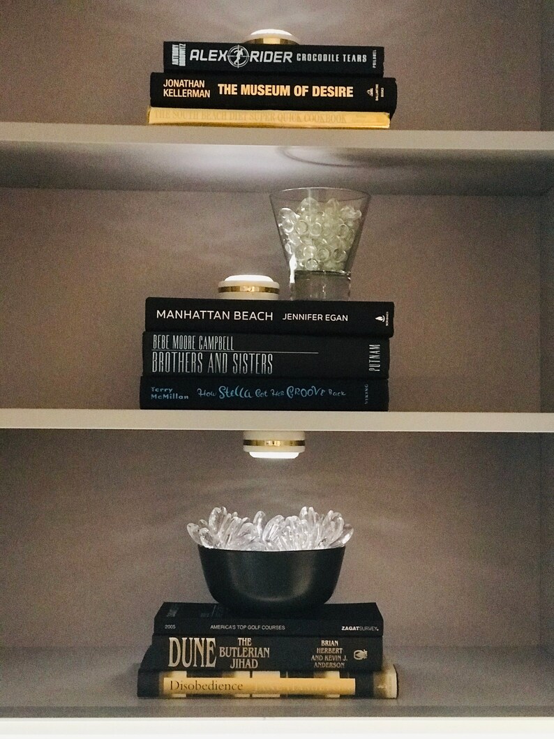 May include: A bookshelf with a variety of books, including "The Museum of Desire" by Jonathan Kellerman, "Manhattan Beach" by Jennifer Egan, and "Brothers and Sisters" by Bebe Moore Campbell. There are also decorative items on the shelves, such as a glass bowl filled with clear glass beads and a black bowl filled with clear glass beads.