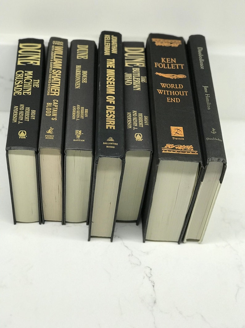 May include: A collection of eight black hardcover books with gold lettering on the spines. The titles include "Dune", "The Machine Crusade", "Captain's Blood", "House Harkonnen", "The Museum of Desire", "The Butlerian Jihad", "World Without End", and "Duobedience".