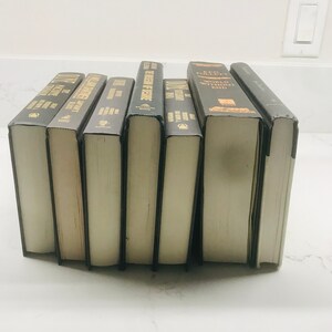 May include: A collection of ten hardcover books with black covers and gold lettering. The titles include "The Pillars of the Earth", "World Without End", "The Historian", "The Alienist", "The Name of the Rose", "The Da Vinci Code", "The Girl with the Dragon Tattoo", "Gone Girl", "The Girl on the Train", and "The Silent Patient".