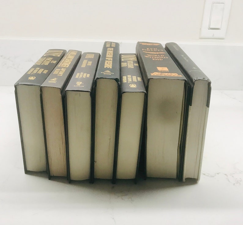 May include: A collection of ten hardcover books with black covers and gold lettering. The titles include "The Pillars of the Earth", "World Without End", "The Historian", "The Alienist", "The Name of the Rose", "The Da Vinci Code", "The Girl with the Dragon Tattoo", "Gone Girl", "The Girl on the Train", and "The Silent Patient".