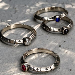 May include: Four silver-toned metal rings featuring a textured band with alternating etched lines and circular indentations. Each ring is set with a single round faceted gemstone in colors including clear, blue, dark purple, and red.
