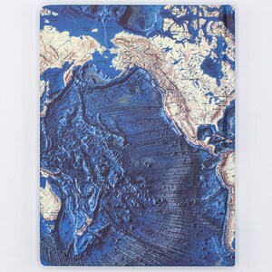 Puede incluir: Un cuaderno azul y blanco con un mapa del mundo con un dise&ntilde;o texturizado en 3D. El mapa muestra la topograf&iacute;a del fondo del oc&eacute;ano con tonos azules profundos y colores azul claro y blanco para las masas terrestres.