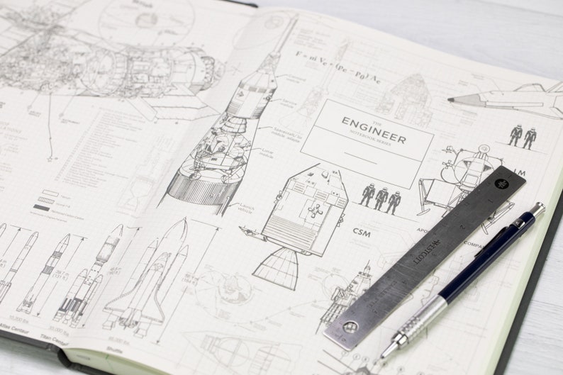 May include: A notebook featuring detailed diagrams of rockets and spacecraft, including a Saturn V. A metal ruler and a dark blue pen are on the pages. The notebook cover reads "THE ENGINEER NOTEBOOK SERIES."