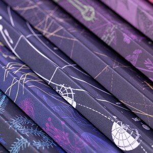 Puede incluir: Una pila de cuadernos de color morado oscuro con diferentes dise&ntilde;os ilustrados en las portadas. Los dise&ntilde;os incluyen una variedad de criaturas marinas, plantas y patrones geom&eacute;tricos.