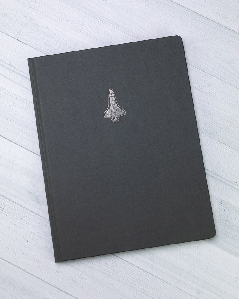 May include: A dark grey notebook featuring a silver space shuttle emblem on the cover. The notebook has a simple, rectangular shape. The background is a light wood grain pattern.