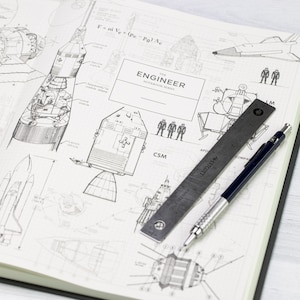 May include: An open notebook featuring detailed diagrams of rockets and spacecraft. A metal ruler and a pen are placed on the open pages. The cover reads "ENGINEER NOTEBOOK SERIES". The pages are white with black ink.
