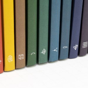 May include: A selection of colourful books with white graphic icons on their spines. The books are in shades of red, yellow, brown, green, blue, and purple. The spines are angled, and the books are on a white surface.