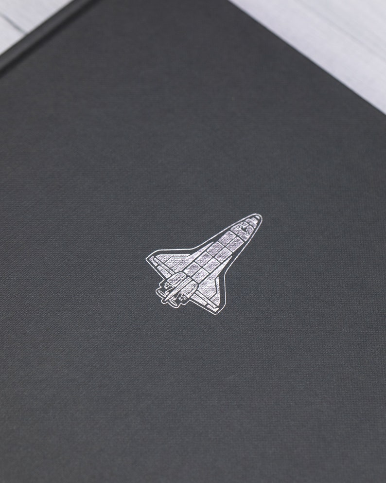 May include: A black notebook featuring a silver illustration of a space shuttle. The image shows a detailed drawing of the spacecraft. The notebook's surface has a textured appearance.