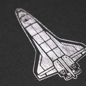 May include: A detailed silver illustration of a space shuttle on a black background. The shuttle design includes wings, body, and engine details. The image has a textured appearance.