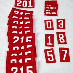 May include: A collection of red fabric patches with white numbers and text. The patches display numbers like 107, 201, 215, 03, 18, 57, and the word "TRAINED". The patches are rectangular.