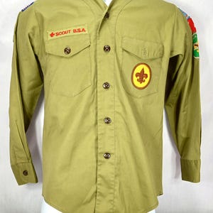 May include: A tan Boy Scout uniform shirt with long sleeves and a button-up front. The shirt has a pocket on each side of the chest and a patch on the right side of the chest that says "SCOUT B.S.A.". The shirt also has a yellow patch with a fleur-de-lis on the right side of the chest.