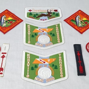 May include: A collection of embroidered patches with various designs and text. Patches include a diamond-shaped orange patch with a Native American figure, a white patch with a deer, and green patches with "Knoxville Tennessee" and "NOAC 2000".