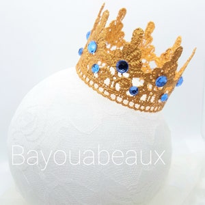 Peut inclure: Une couronne dor&eacute;e orn&eacute;e de pierres bleues, pos&eacute;e sur une surface blanche. La couronne est en dentelle et arbore un design d&eacute;licat.