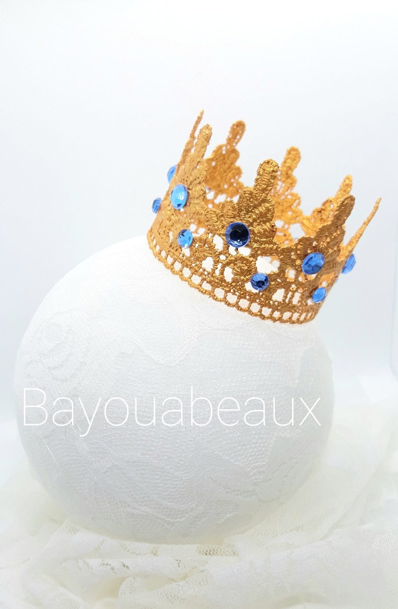 Peut inclure: Une couronne dor&eacute;e orn&eacute;e de pierres bleues, pos&eacute;e sur une surface blanche. La couronne est en dentelle et arbore un design d&eacute;licat.