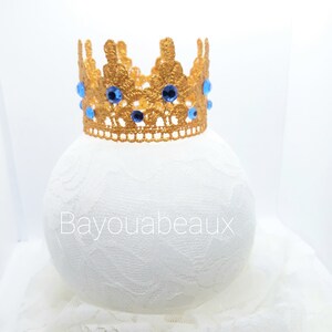 Peut inclure: Une couronne dor&eacute;e avec des strass bleus. La couronne est en dentelle crochet&eacute;e et repose sur un fond de dentelle blanche.