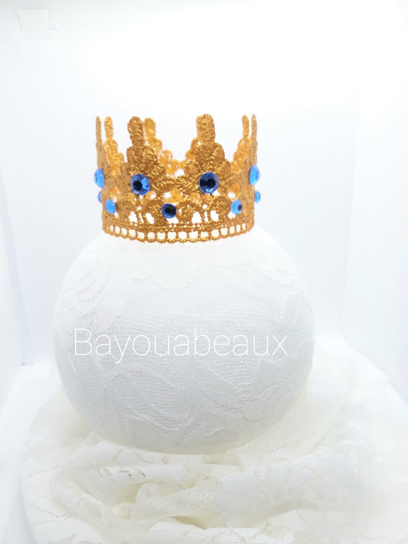 Peut inclure: Une couronne dor&eacute;e avec des strass bleus. La couronne est en dentelle crochet&eacute;e et repose sur un fond de dentelle blanche.