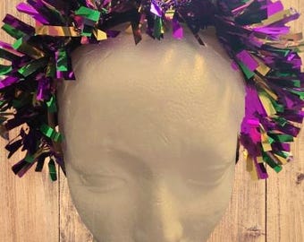 May include: Purple, green, and gold metallic streamer headband. The headband is made of thin, shiny strips of material that are gathered together to form a crown-like shape.