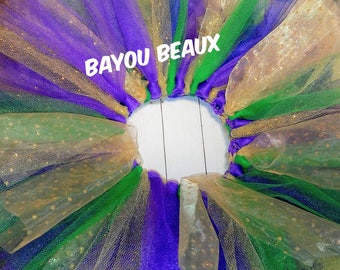 May include: A multi-layered tulle skirt in gold, green, and purple with gold glitter accents. The skirt has a gathered waistband and the text "BAYOU BEAUX" is printed on the waistband.