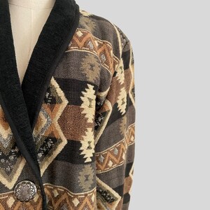 May include: A tapestry-style jacket with a Southwestern geometric pattern in shades of brown, beige, and black. The jacket features a black collar and trim, and a silver-tone button. The overall design is reminiscent of vintage or bohemian fashion.