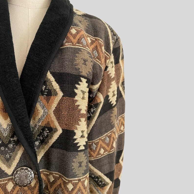 May include: A tapestry-style jacket with a Southwestern geometric pattern in shades of brown, beige, and black. The jacket features a black collar and trim, and a silver-tone button. The overall design is reminiscent of vintage or bohemian fashion.
