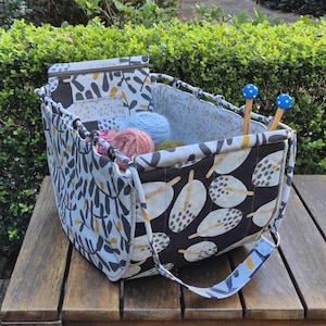 Puede incluir: Una cesta de almacenamiento rectangular de tela con un patrón de hojas en blanco y negro en el exterior y un interior azul claro. La cesta incluye una solapa superior con cremallera, dos asas azules con lunares blancos y contiene ovillos de lana.