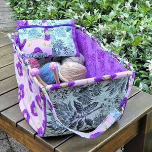 Puede incluir: Una bolsa de tela para proyectos de tejido con un estampado floral en tonos morados, grises y blancos. La bolsa contiene lana y agujas de tejer. Una bolsa a juego está dentro. La bolsa tiene un asa y está sobre una superficie de madera.