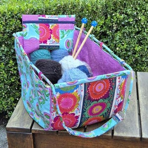 May include: A rectangular fabric storage basket with a light blue exterior featuring a colourful floral pattern in pink, orange, and green. The interior is lined with purple fabric. It contains several balls of yarn and two wooden knitting needles.