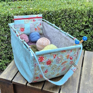 May include: A turquoise fabric knitting bag with a floral design and a matching pouch. The bag holds several balls of yarn in shades of pink, blue, and yellow. A pair of blue and white knitting needles are visible.